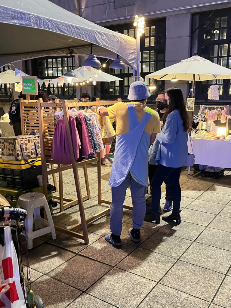 夜間市集裡的攤架：梯型框架掛著童裝，頂上吊著燈，角落掛著綠色招牌