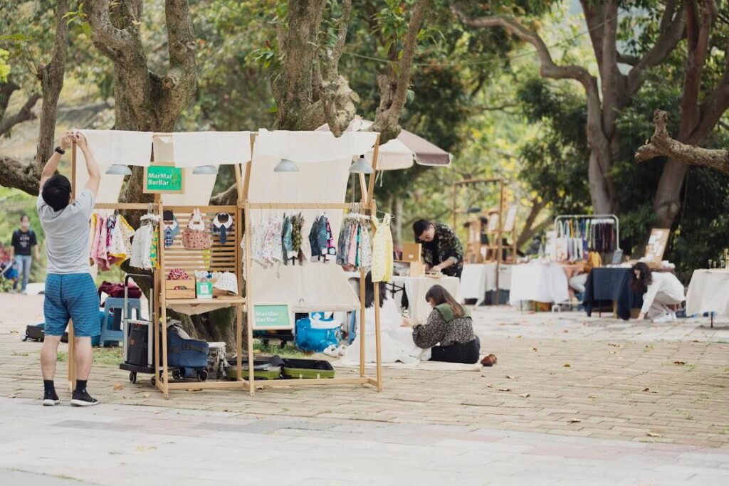 戶外市集裡的 Marmarbarbar 攤位：木框架掛滿童衣，頂上撐著白布，掛著綠色招牌