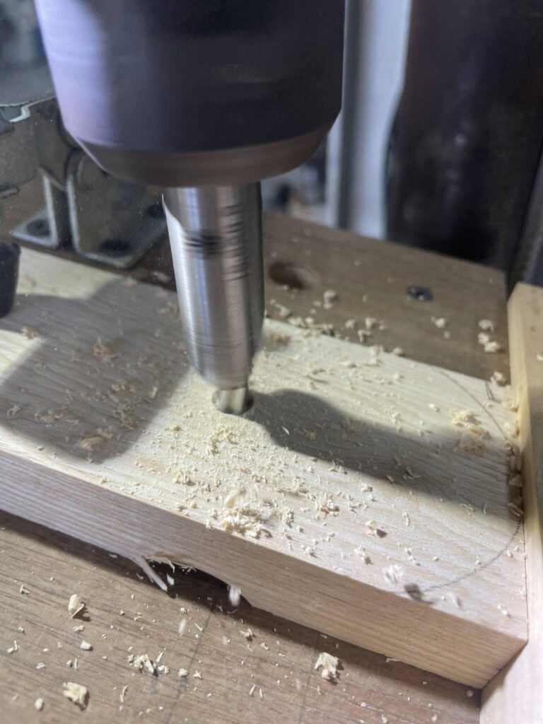 鑽床正在鑽孔，工件壓在自製的木質治具板上，旁有金屬固定件與木屑
